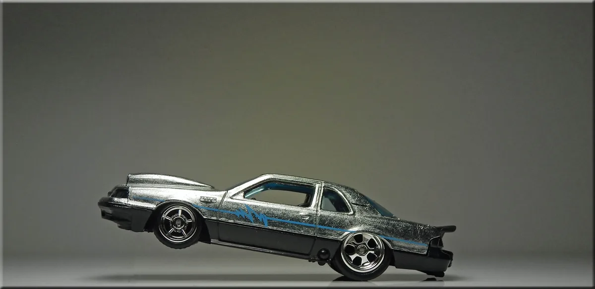 A silver and black Hot Wheels die-cast model car with blue racing stripes is photographed in profile against a plain gray…