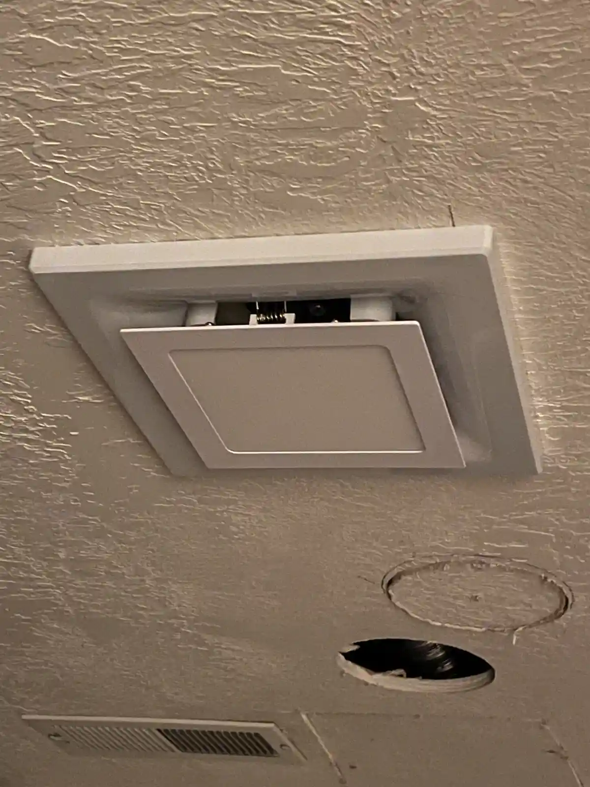 A close-up photo shows a ceiling with a square white light fixture partially open, revealing its internal wiring and…