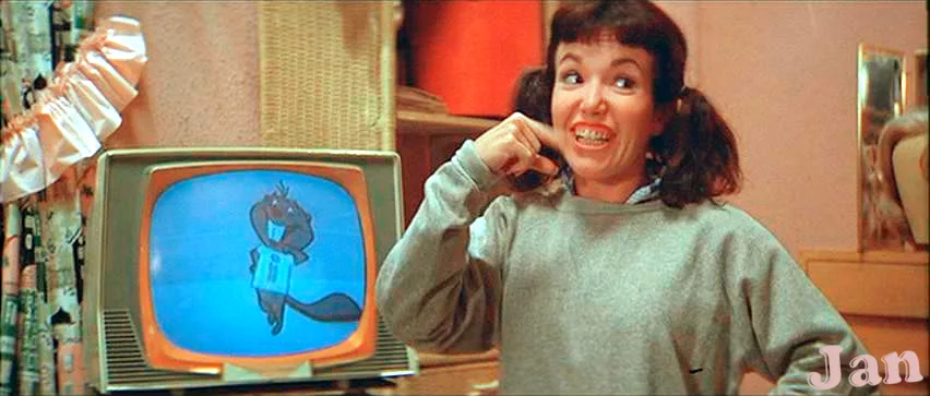 A woman with pigtails and a grey sweatshirt stands beside a vintage television, pointing at it with a wide, excited grin…