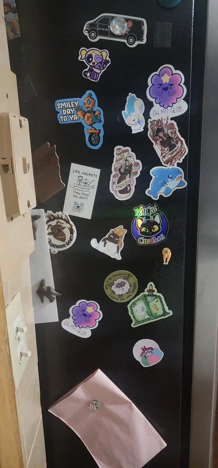 A black refrigerator door is covered with numerous colorful magnets and stickers, including cartoon characters like the…