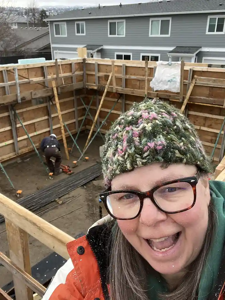 A woman wearing a colorful knit hat and glasses smiles broadly at the camera while standing on a construction site, with…
