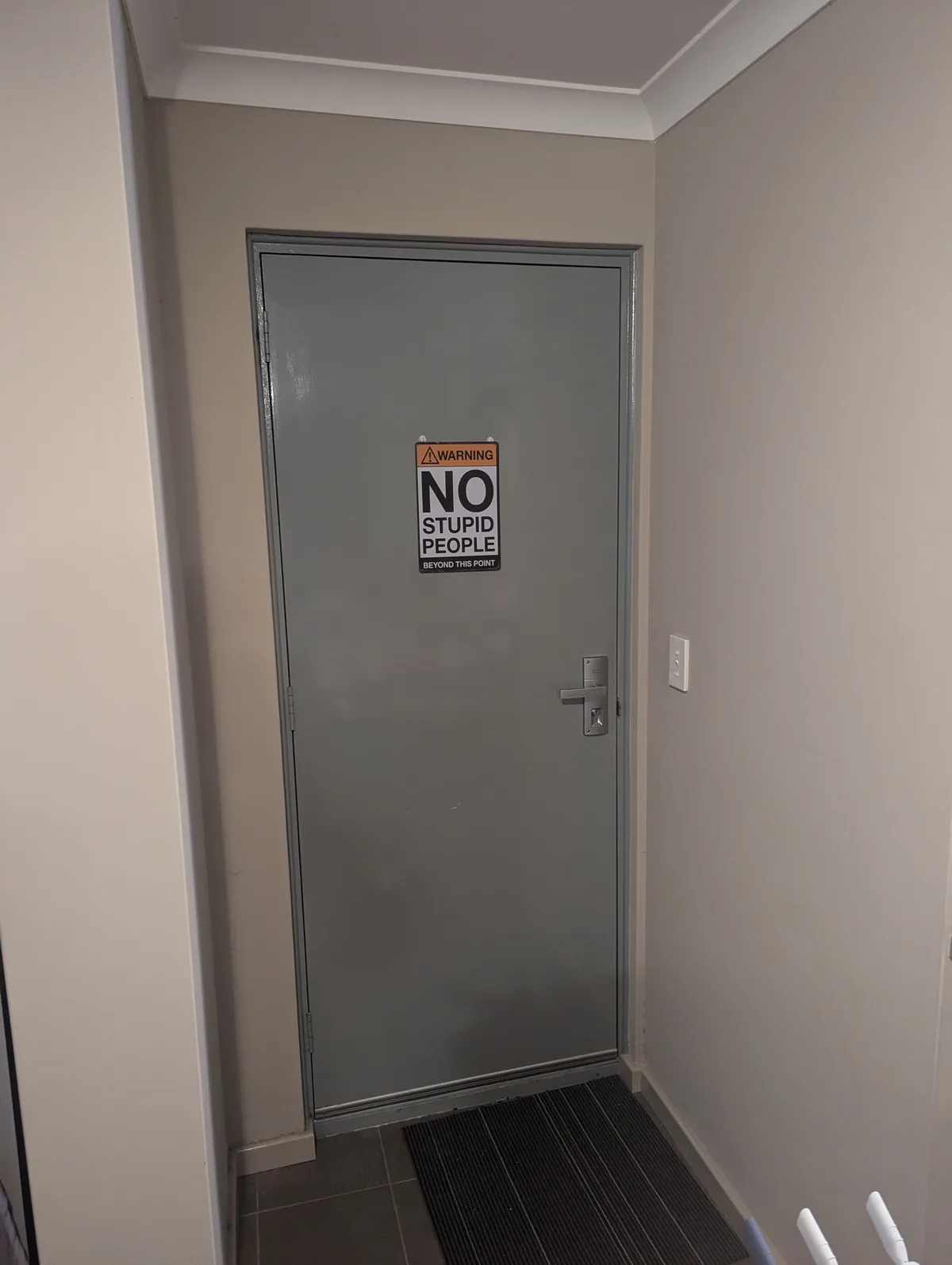 A plain gray door in a hallway has a humorous warning sign taped to it that reads 'WARNING NO STUPID PEOPLE BEYOND THIS…