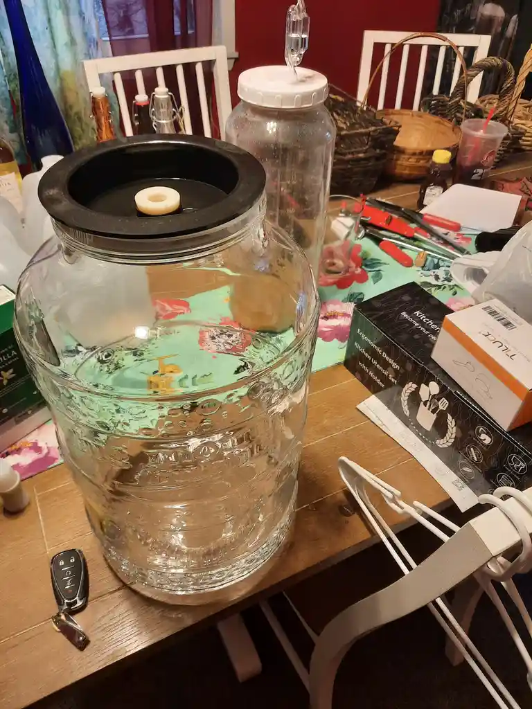 A large glass fermentation keg filled with clear water sits on a cluttered wooden table, topped with a black rubber gasket…