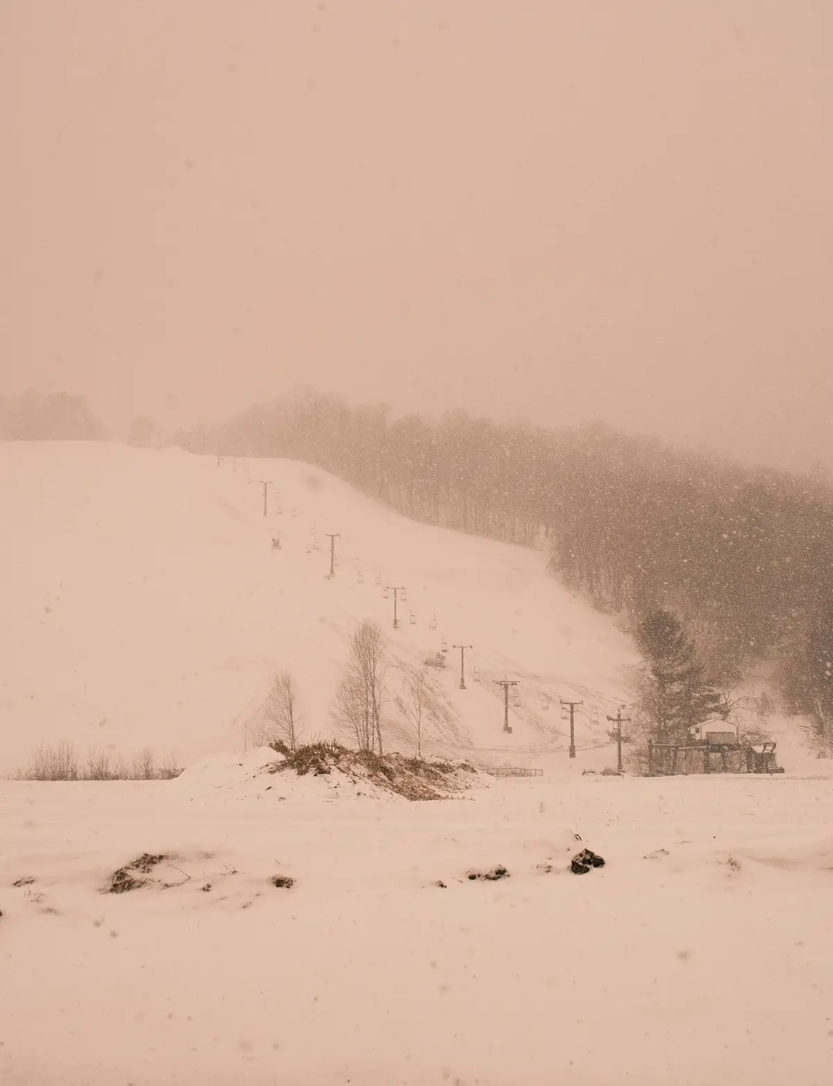 A snowy, blizzard-covered ski slope with a chairlift visible through falling snow, featuring a small building and bare trees…
