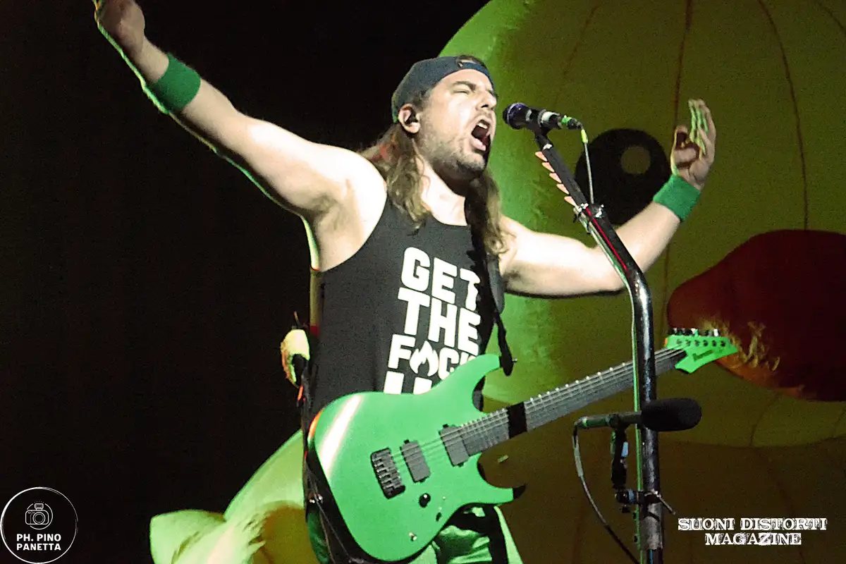 A male musician with long hair and a backwards cap is performing on stage, arms raised, singing into a microphone while…