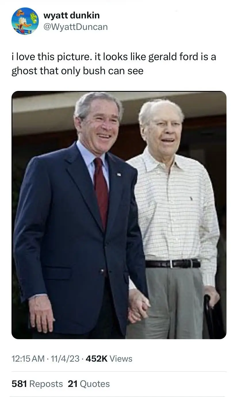 The image shows two elderly men standing side by side, one in a dark suit and red tie, the other in a light-colored…