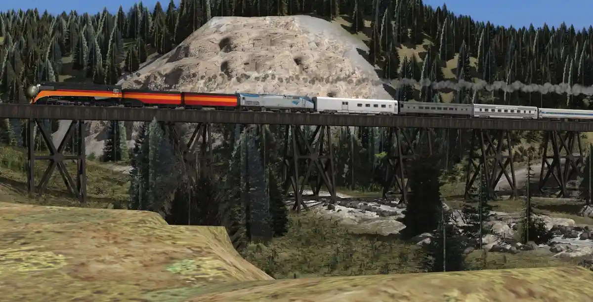 A long passenger train with a black and orange locomotive and silver passenger cars travels across a wooden trestle bridge…