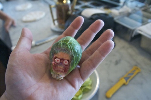 a person holds a small brussel sprout in their palm, which has been crudely carved or painted to resemble a grotesque, angry…