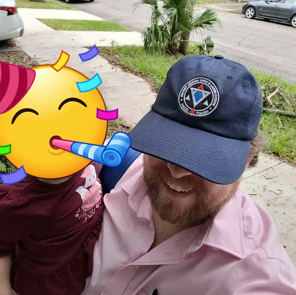 A man with a beard and a blue baseball cap featuring a 'Secret Jewish Space Laser Corps' logo smiles while holding a child…