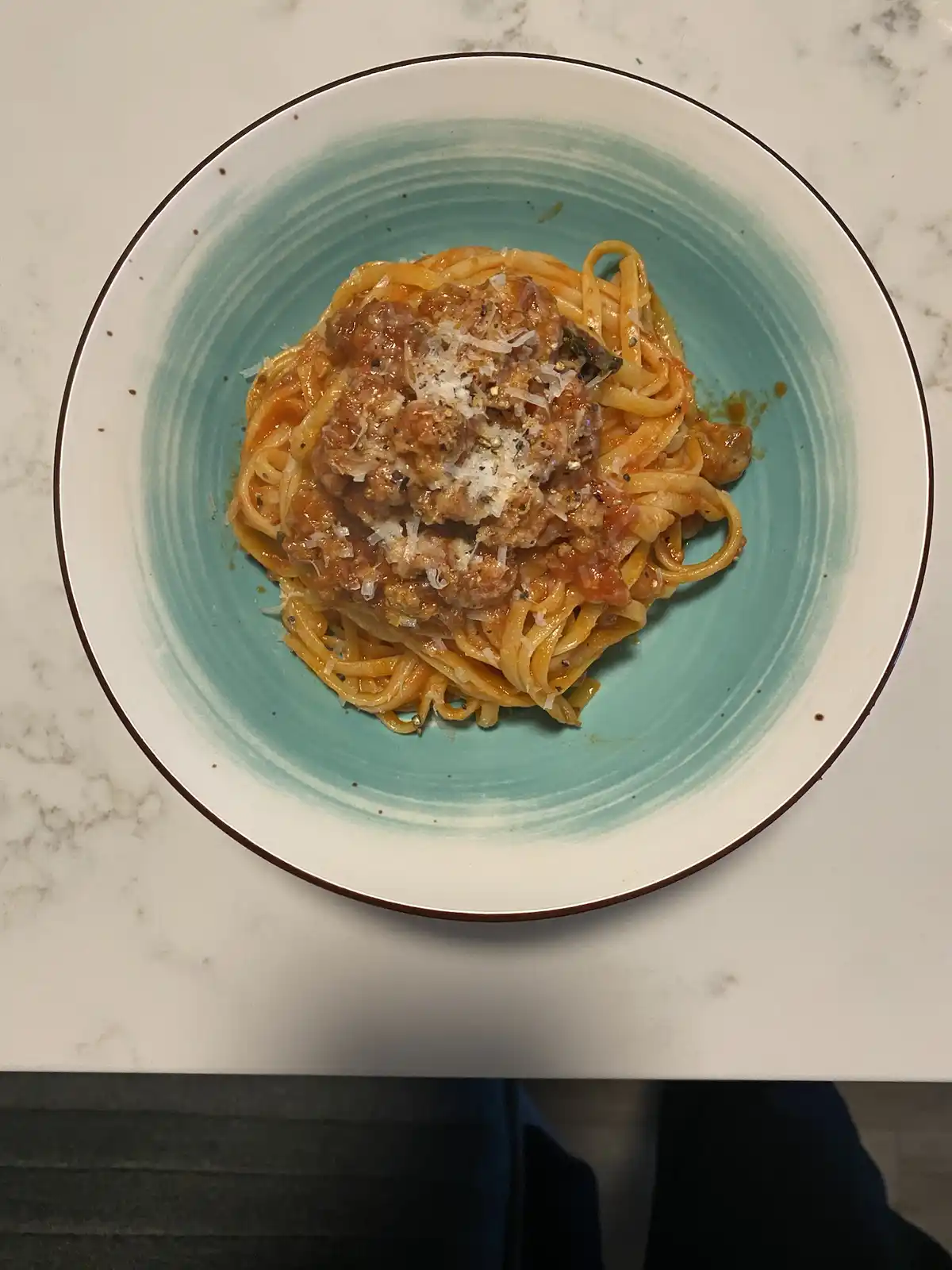 A top-down photo shows a bowl of spaghetti with meat sauce, grated cheese, and black pepper, served on a light blue ceramic…