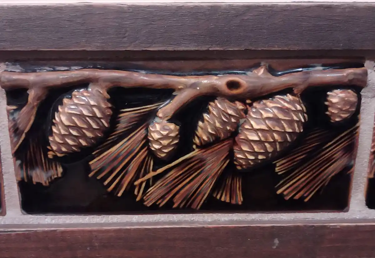 A close-up of a dark brown ceramic tile with a raised relief design of pinecones and pine needles hanging from a branch, set…