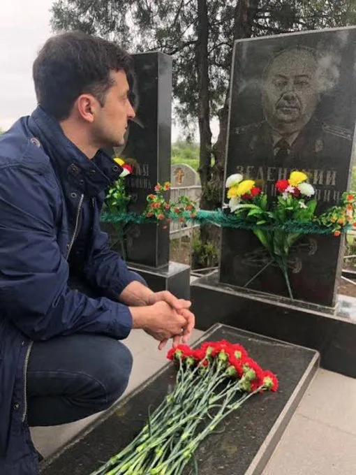 A man in a dark blue jacket kneels before a grave marker featuring a portrait of a man with the name 'ЗЕЛЕНСЬКИЙ'…