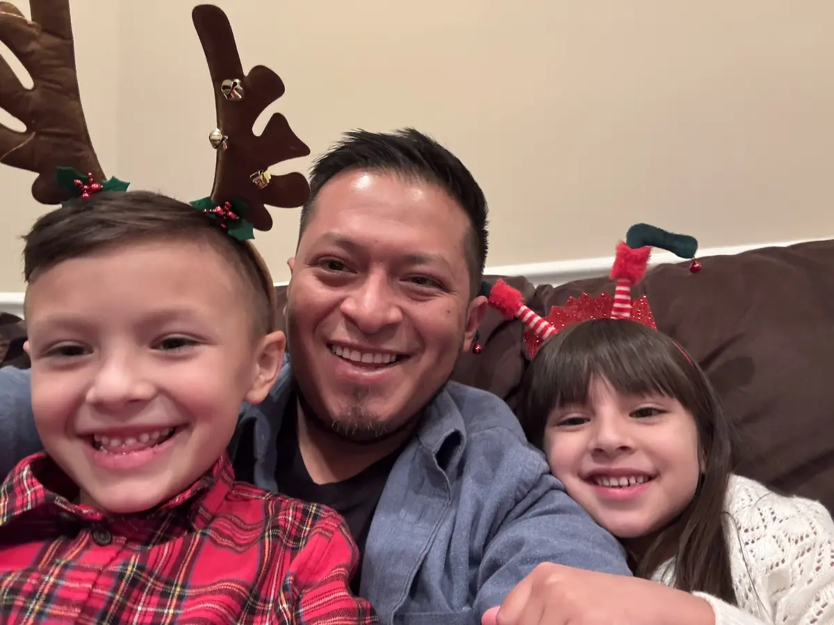 A smiling man with two children sits on a couch, all wearing festive holiday headbands. The boy on the left wears brown…