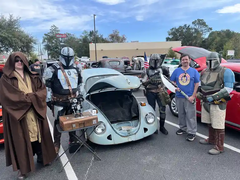 Five men in Star Wars costumes — a Jedi, three Mandalorians, and a man in a blue Star Wars t-shirt — stand around a light…