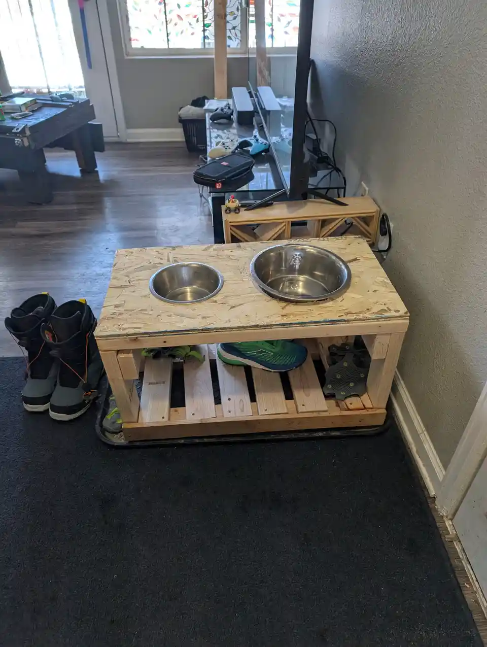 A DIY wooden table with two stainless steel dog bowls on top and shoes stored underneath, placed in a living room near a TV…