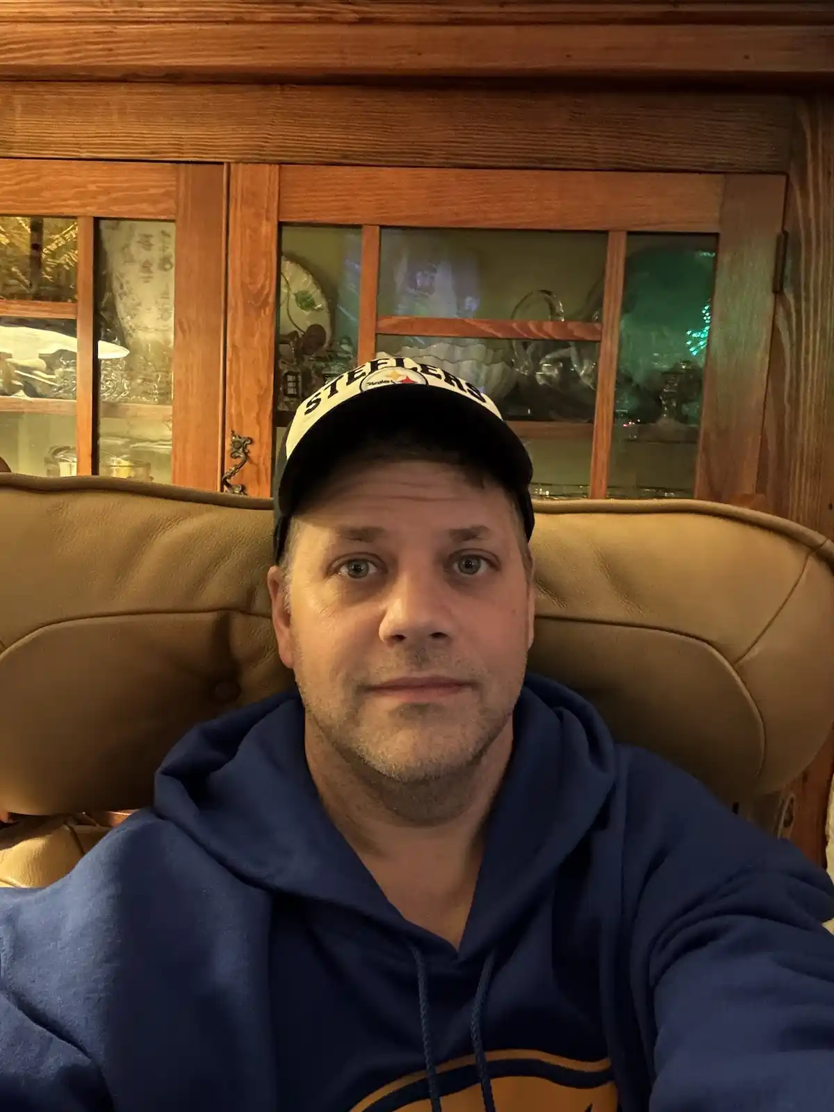 A man wearing a Pittsburgh Steelers baseball cap and a blue hoodie sits in a tan leather chair, looking directly at the…