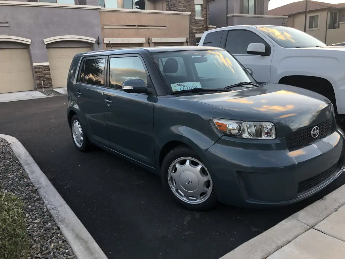 A dark green, boxy Scion xB is parked in a residential driveway next to a white truck, with houses and garages in the…