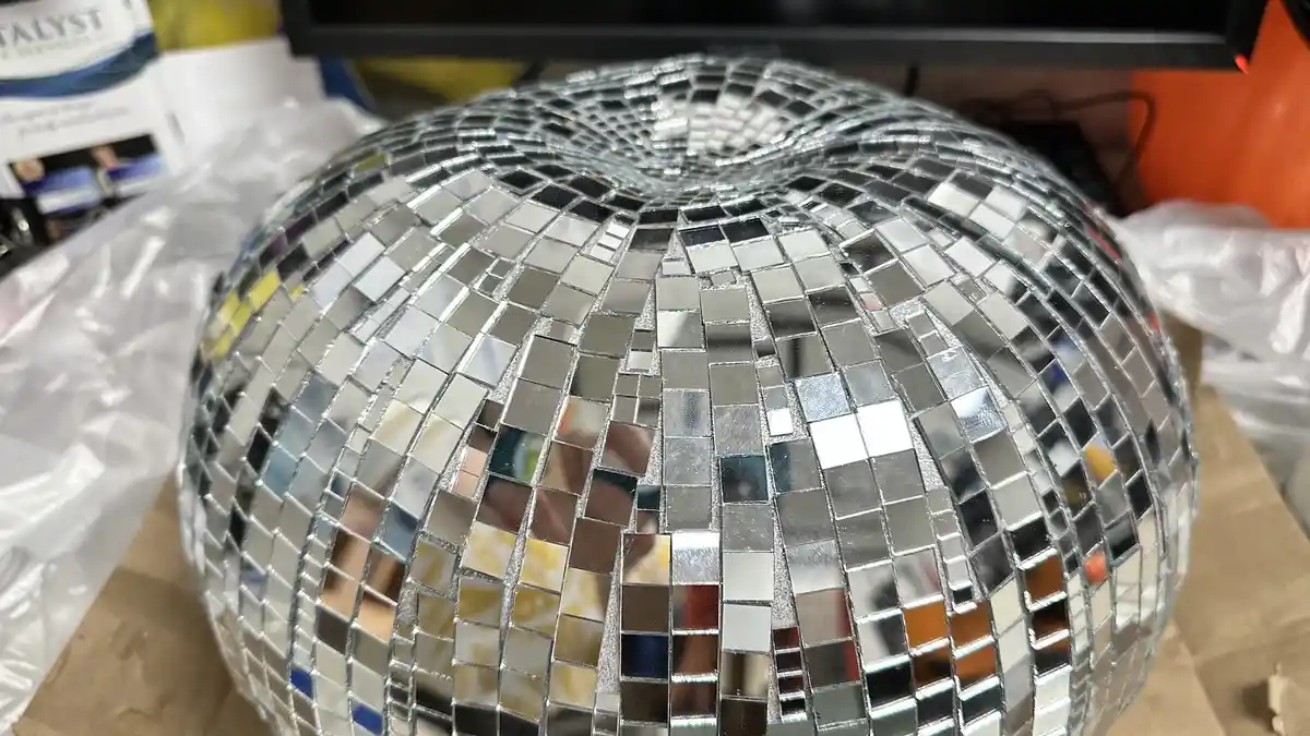 A close-up photo shows a large, spherical disco ball made of many small, square, mirrored tiles reflecting light and…