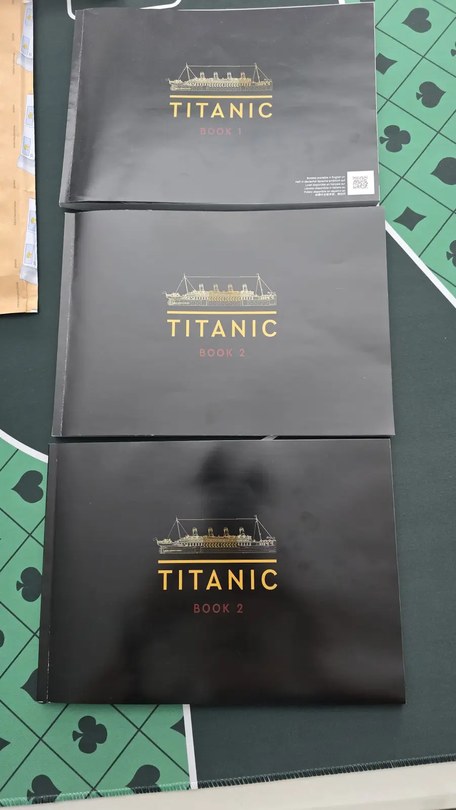 Three black books titled 'Titanic Book 1', 'Titanic Book 2', and 'Titanic Book 2' are stacked on a green felt surface with…