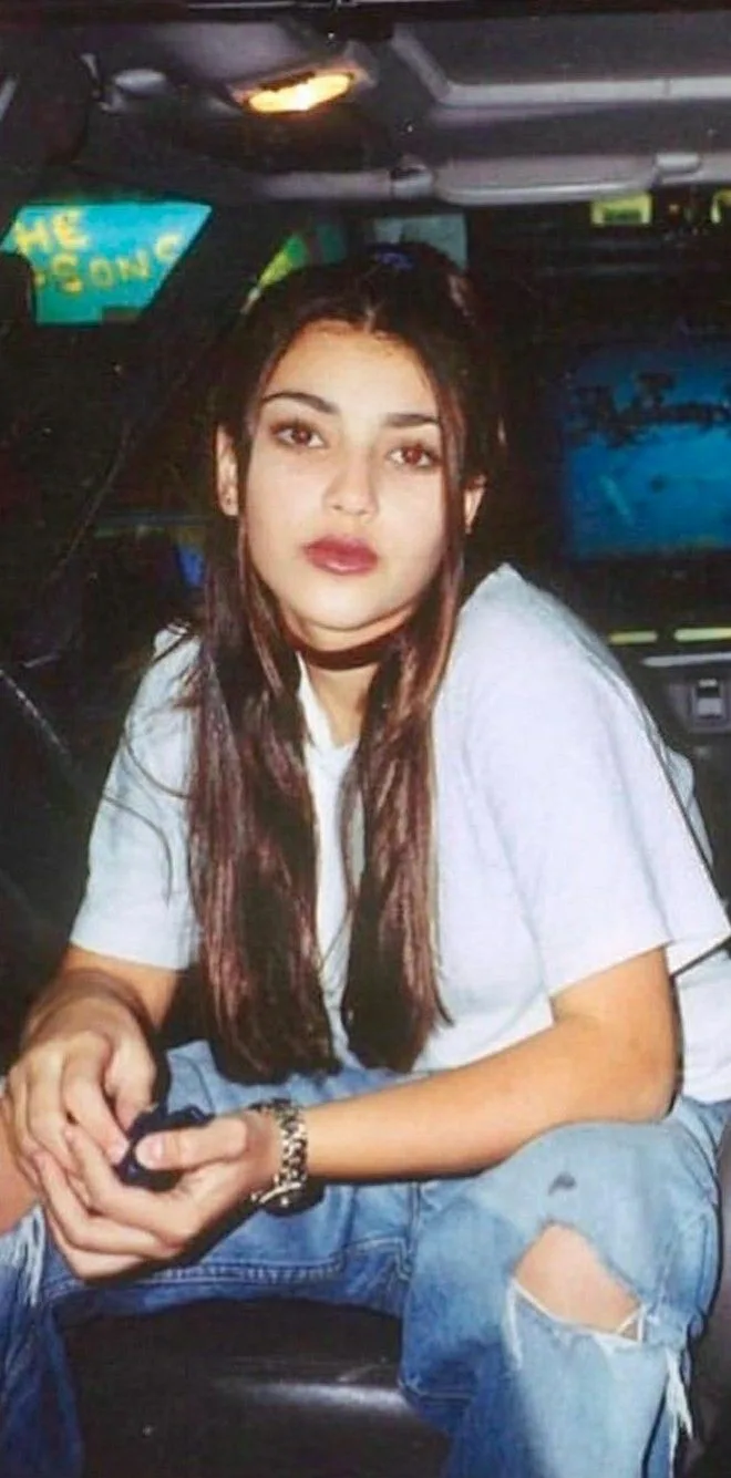 A young woman with long brown hair sits inside a vehicle, wearing a light blue t-shirt and ripped jeans, looking directly at…