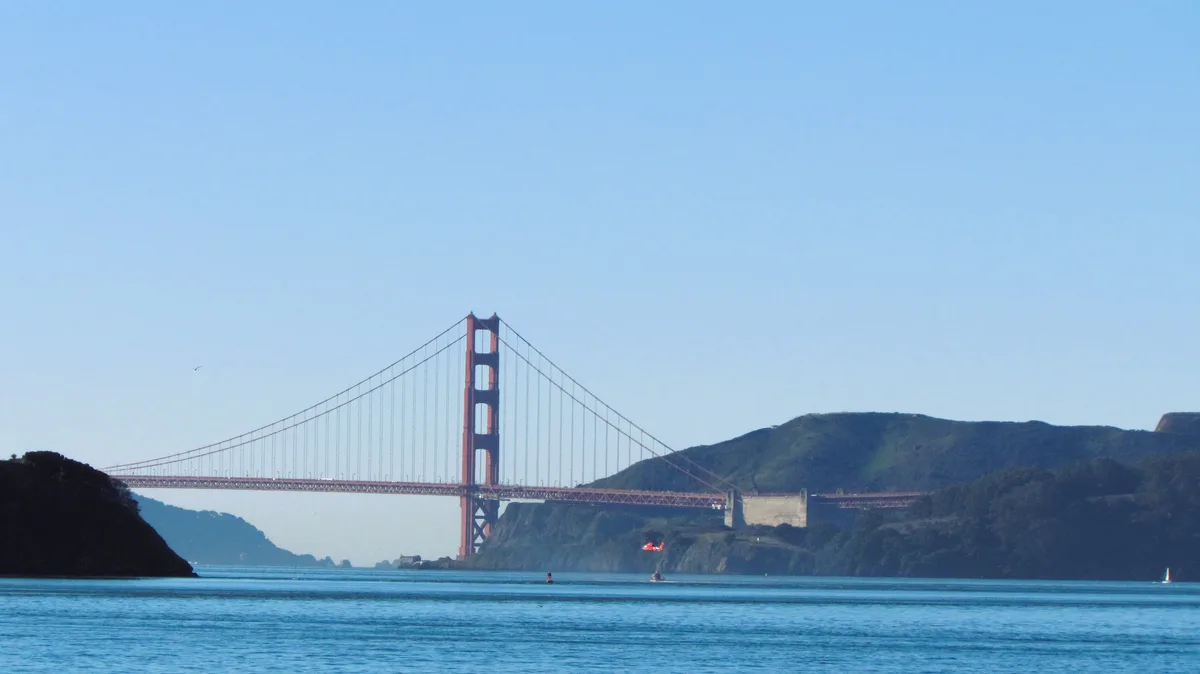 A scenic view of the Golden Gate Bridge under a clear blue sky, with water in the foreground and hills in the background. A…