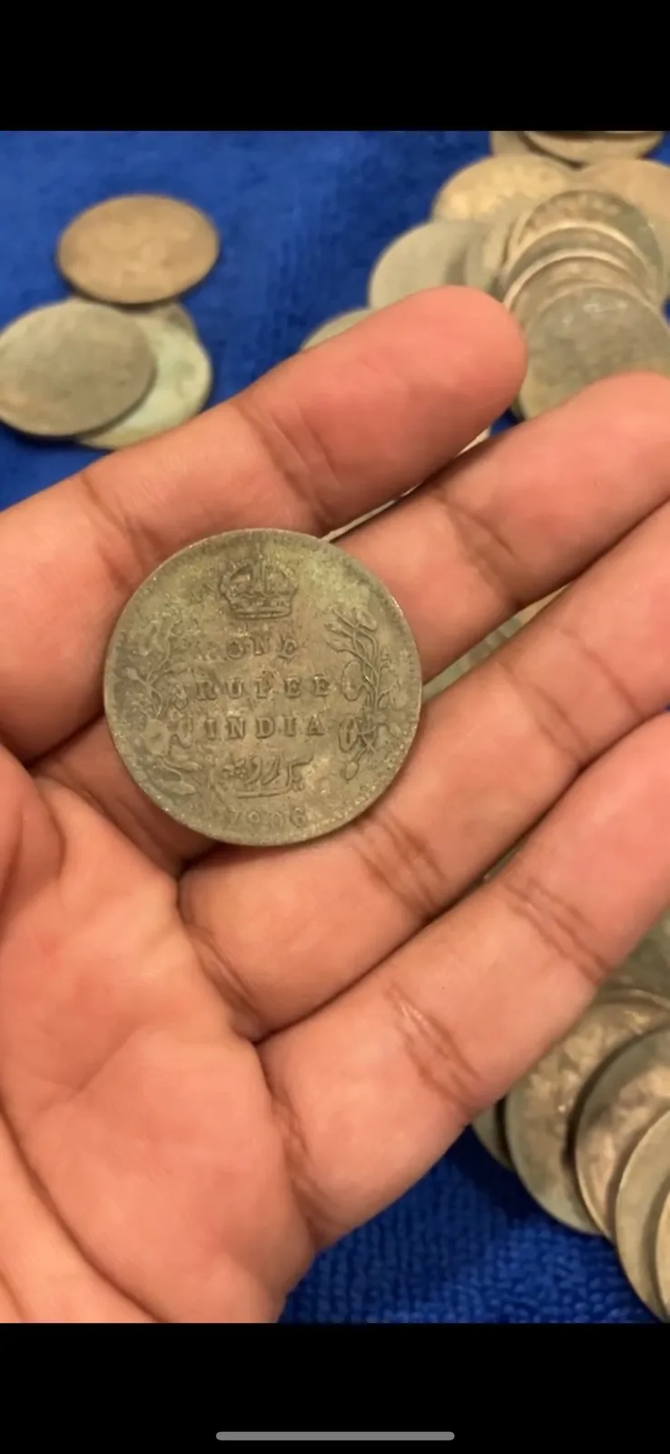 A hand holds a tarnished silver coin inscribed 'ONE RUPEE INDIA 1906' with a crown above, surrounded by other similar coins…