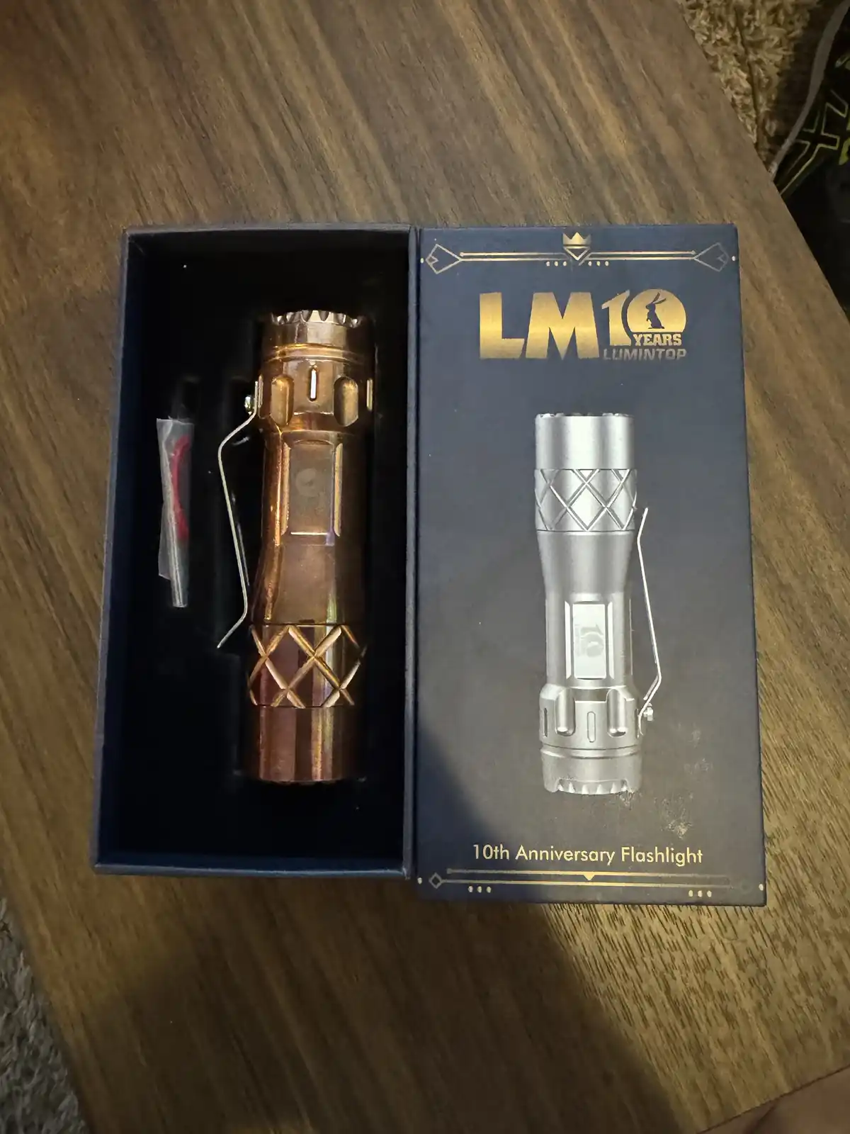 A copper-colored Lumintop LM10 10th anniversary flashlight sits in its open blue box on a wooden table, next to the box's…