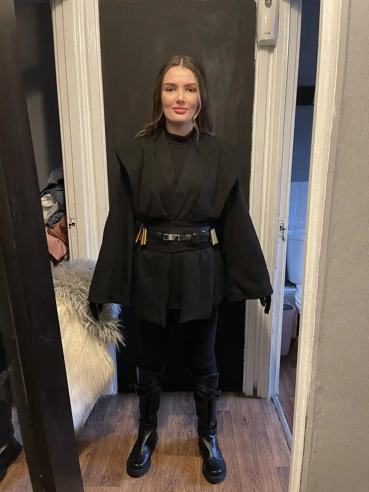 A woman stands in a doorway wearing a black Jedi-style robe, black leggings, and tall black boots with straps, posing for…