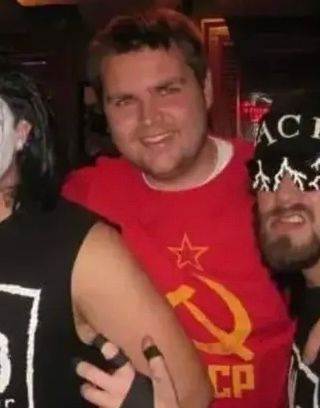 A man in a red t-shirt with a yellow hammer and sickle and star logo smiles while flanked by two other men in dark clothing…
