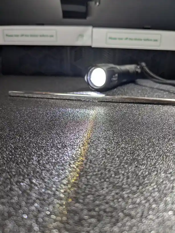 A close-up photo shows a black flashlight and a metal tool resting on a textured black surface, with a bright light…