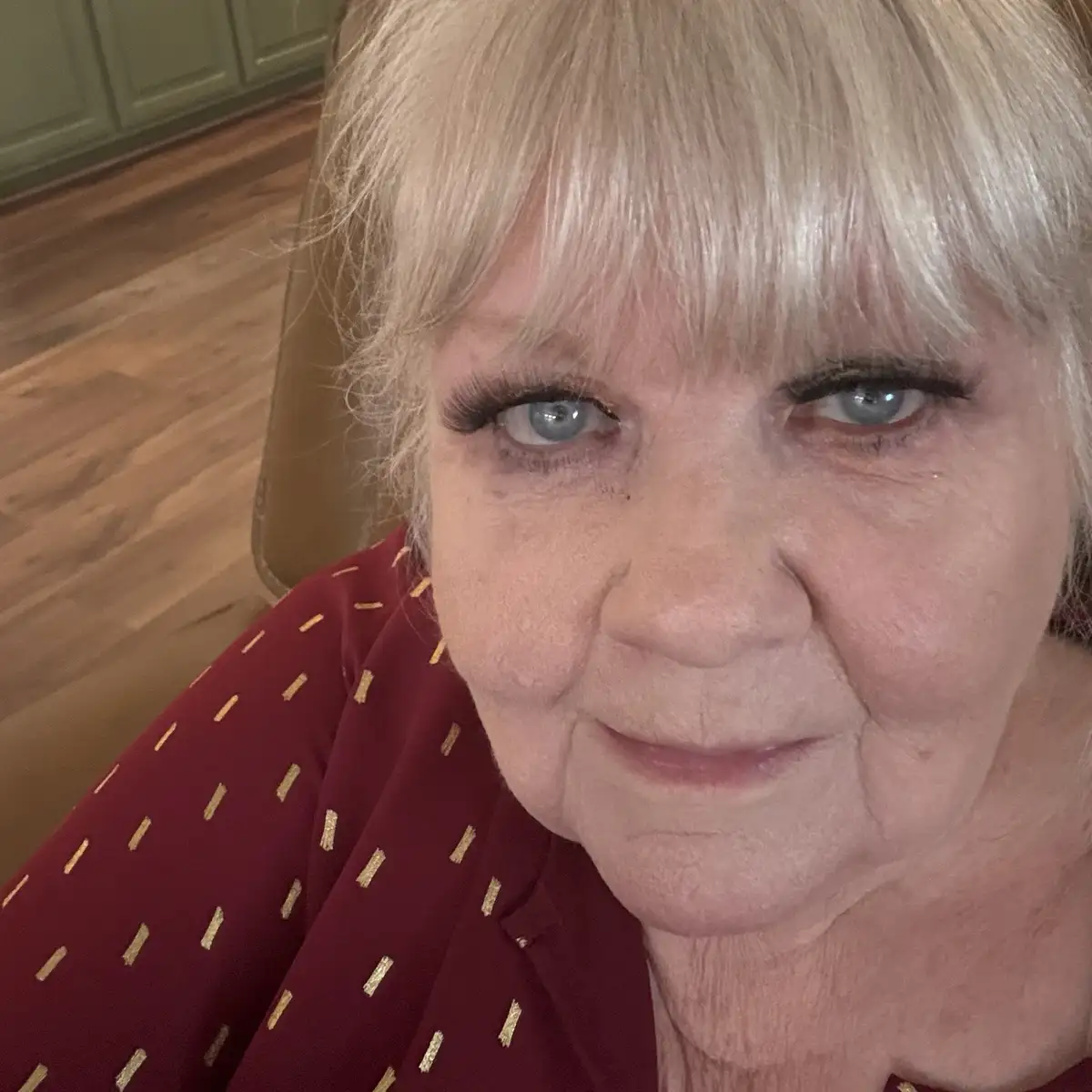A close-up selfie of a woman with blonde hair and blue eyes, wearing a red shirt with gold patterns, looking directly at the…