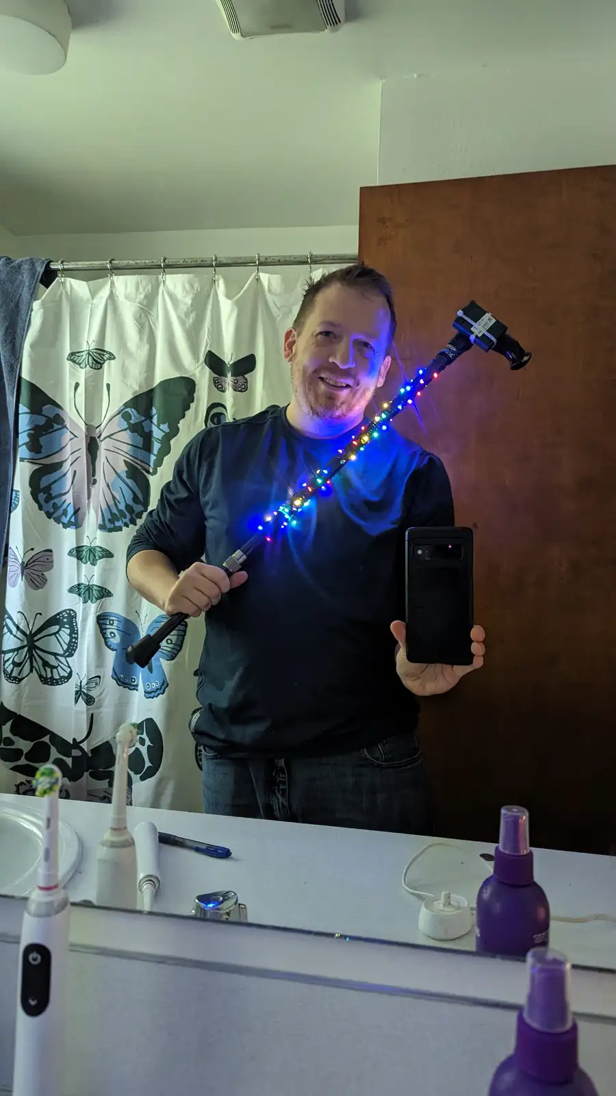 A man in a bathroom mirror holds a smartphone in one hand and a selfie stick with colorful LED lights attached to it in the…