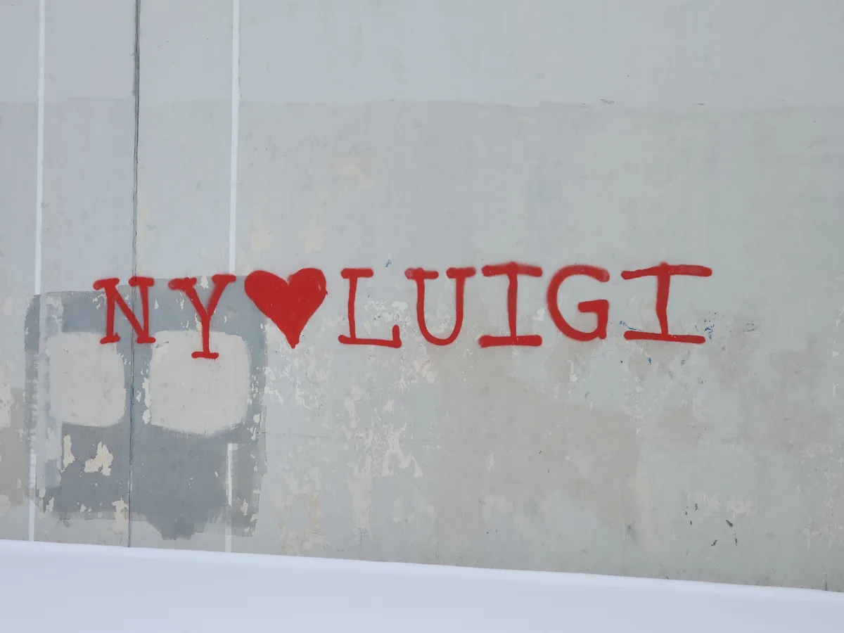 Red spray-painted graffiti on a weathered gray concrete wall reads 'NY❤️LUIGI', with a heart symbol between the letters. The…