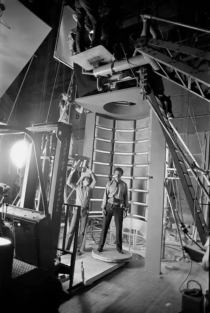 A black-and-white photo shows a man standing on a circular platform in a complex film studio setup with rigging, lights, and…
