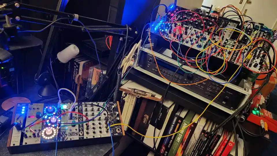 A cluttered music studio setup featuring multiple synthesizers and audio equipment, all interconnected by a chaotic tangle…