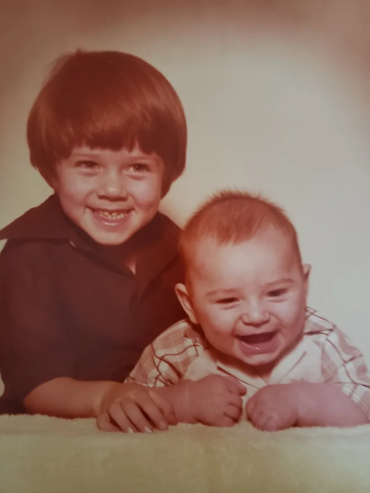 A vintage, sepia-toned photo shows an older boy with a toothy grin standing behind a younger baby who is laughing joyfully…