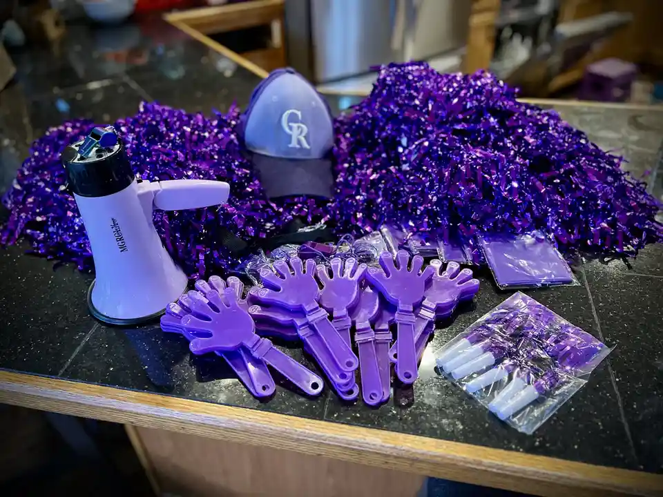 A kitchen counter is covered in purple sports fan gear including a megaphone, a baseball cap with a 'CR' logo, large piles…