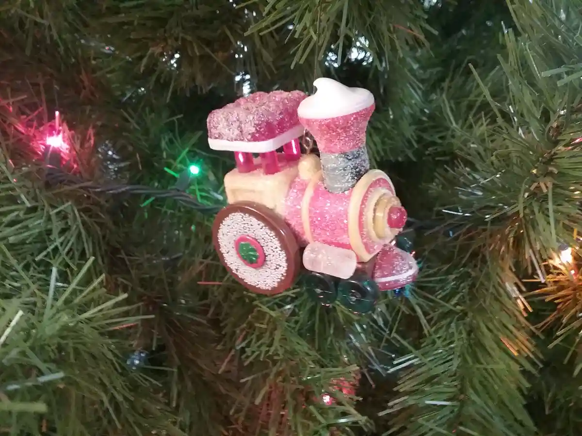 A sparkly pink train ornament hangs on a green Christmas tree with colorful lights, with the text 'i'm not a train, i'm a…