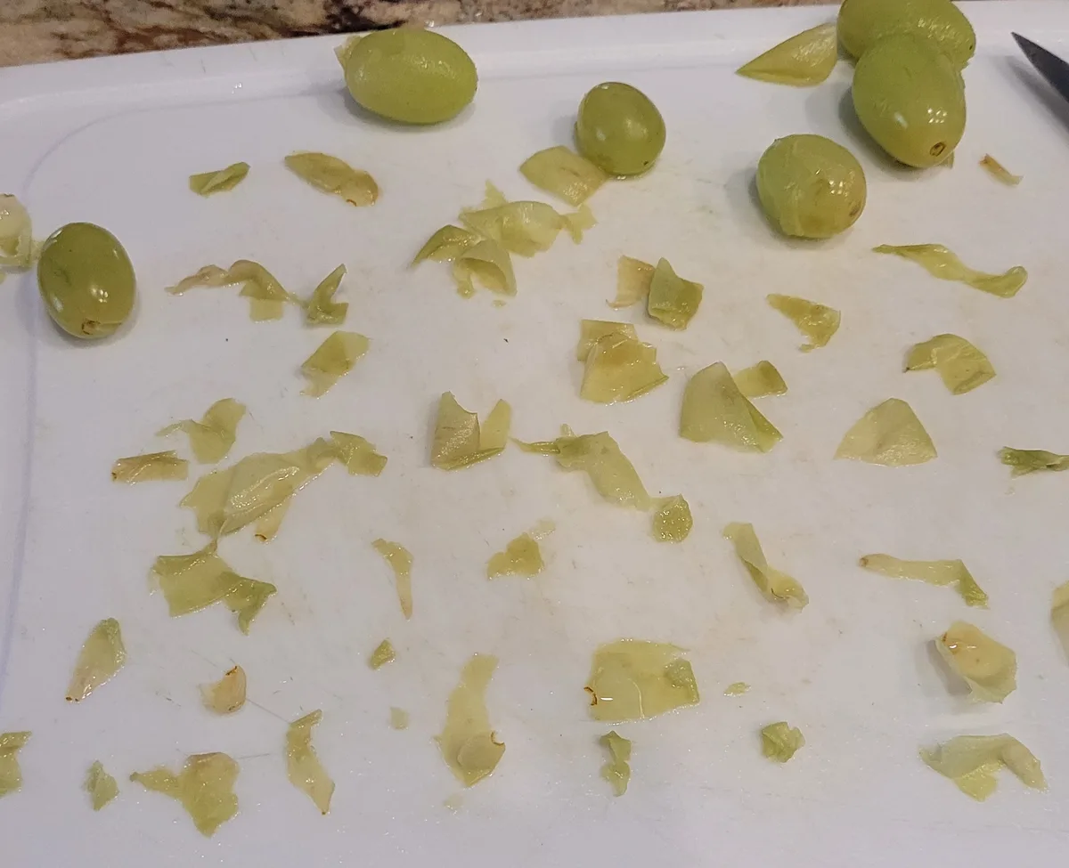 A white cutting board has several green olives scattered across it, some whole and some partially peeled, surrounded by a…
