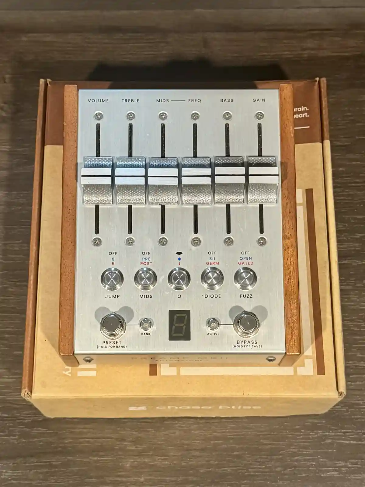 A silver Chase Bliss Preamp MKII guitar effects pedal sits in its cardboard box on a wooden surface. The pedal has six large…