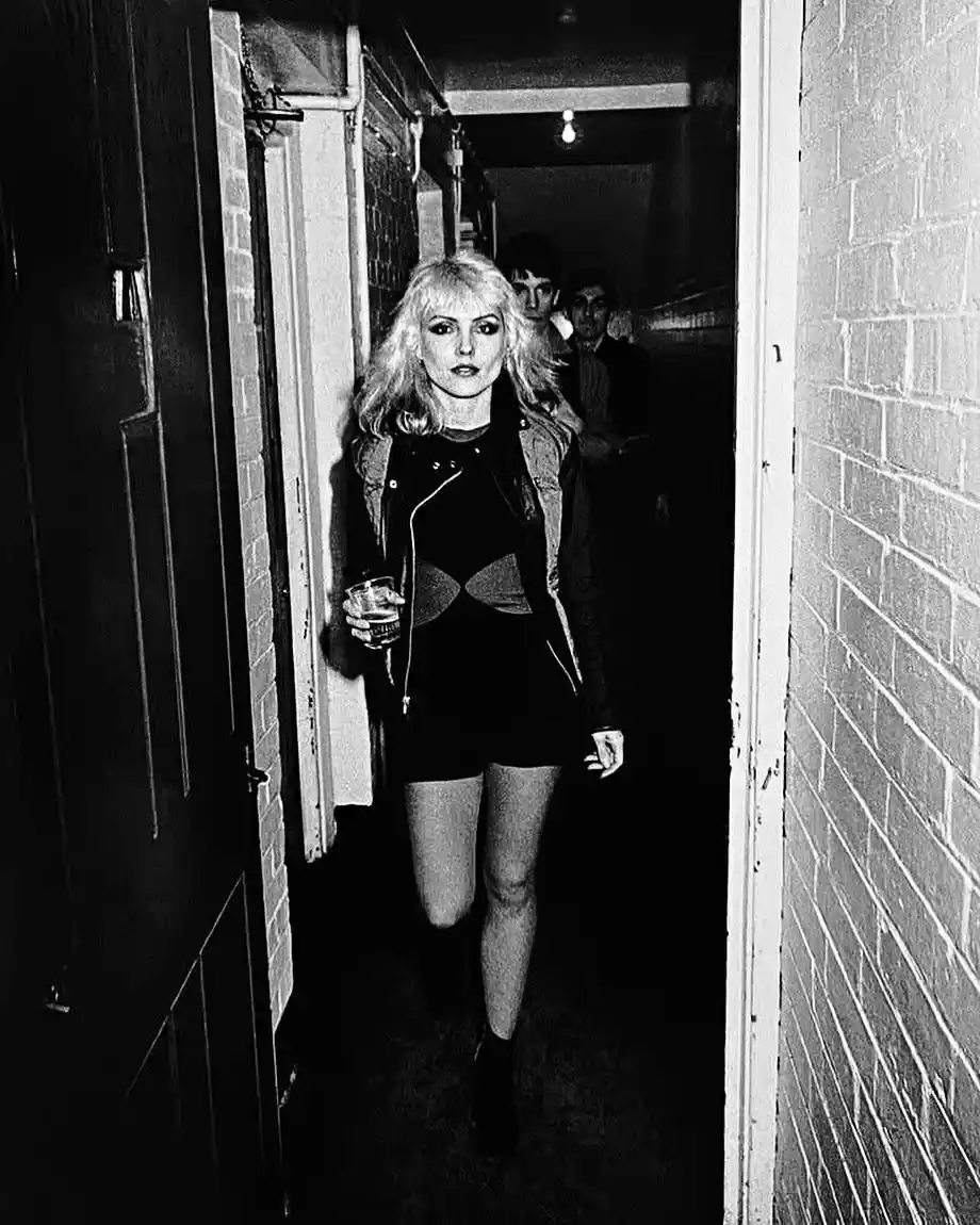A black-and-white photo shows a woman with blonde hair, wearing a leather jacket and holding a drink, walking down a narrow…