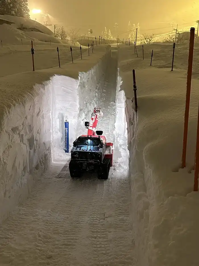A red and black snowplow vehicle is driving through a narrow, freshly carved tunnel in deep snow, with high snow walls on…