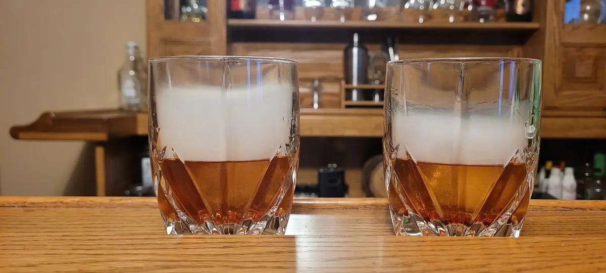 Two identical cut-glass tumblers filled with amber-colored whiskey topped with a thick white foam sit side-by-side on a…