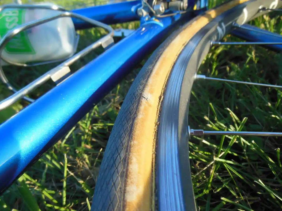 A close-up photo of a blue bicycle lying on grass, showing a severely worn and cracked tire sidewall. A water bottle is…