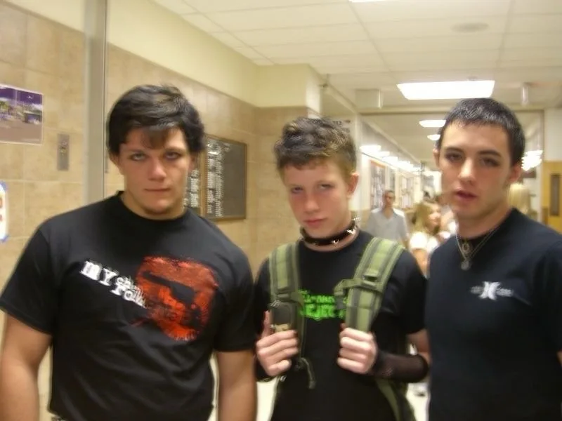 Three teenage boys stand in a school hallway, looking at the camera with serious expressions. The boy on the left wears a…