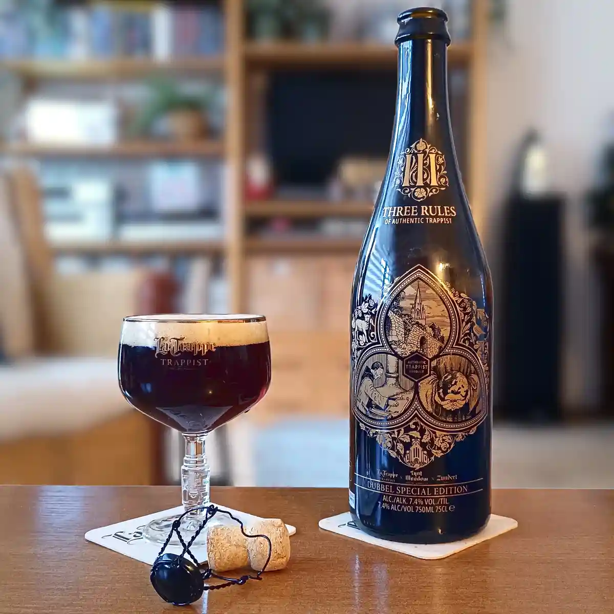 A dark blue bottle labeled 'Three Rules of Authentic Trappist' sits next to a glass of dark beer on a wooden table, with a…