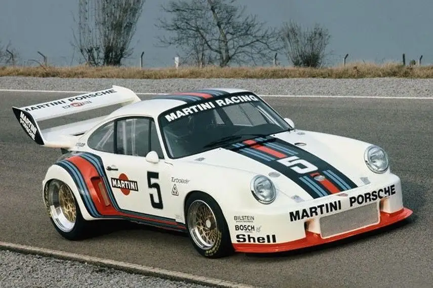 A white Porsche 934 race car with Martini Racing livery, featuring blue, red, and black stripes, the number 5, and sponsor…