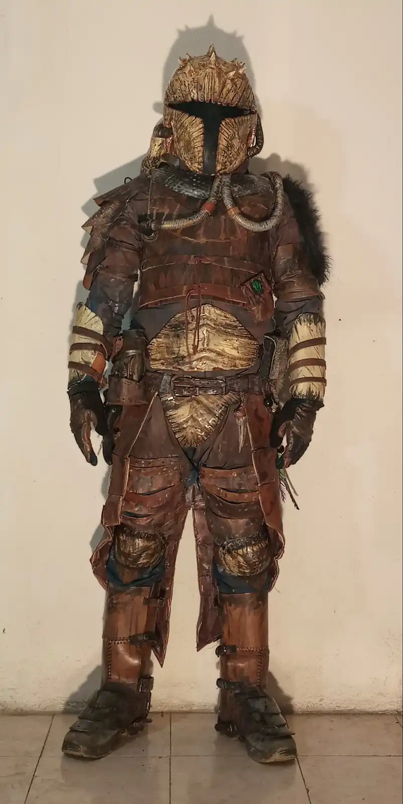 A person stands full-body in a heavily worn, brown leather and metal armor costume resembling a Mandalorian, with a spiked…