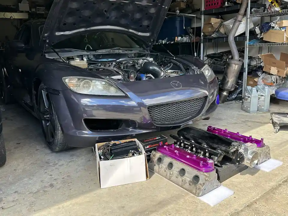 A dark purple Mazda RX-8 sits in a cluttered garage with its hood open, revealing the engine bay. In front of the car, on…