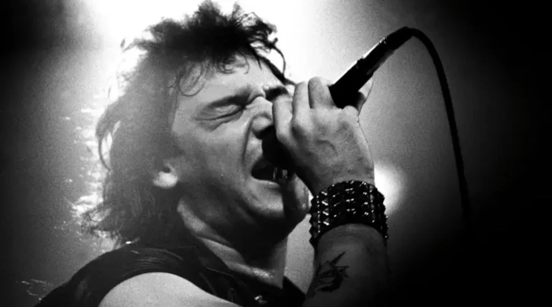 A black and white photo of a male rock singer with long hair, eyes closed, and mouth wide open as he screams into a…